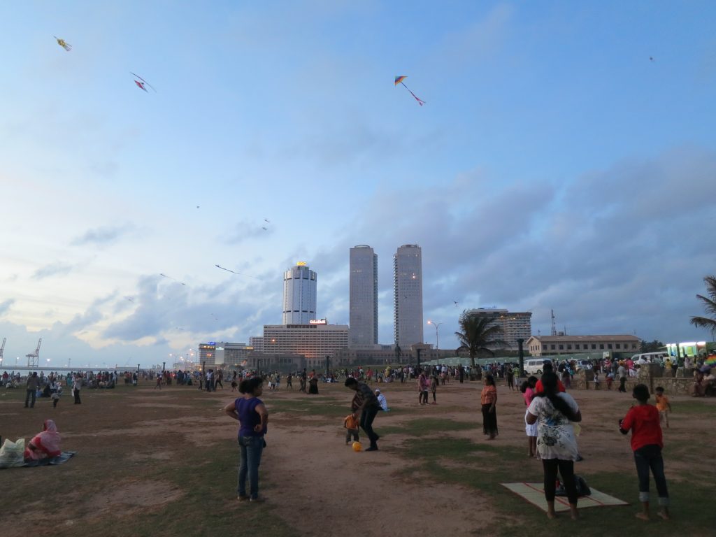 Families and kites on the lawn of Galle Face Green in the evening, with Colombo's World Trade Center towers behind