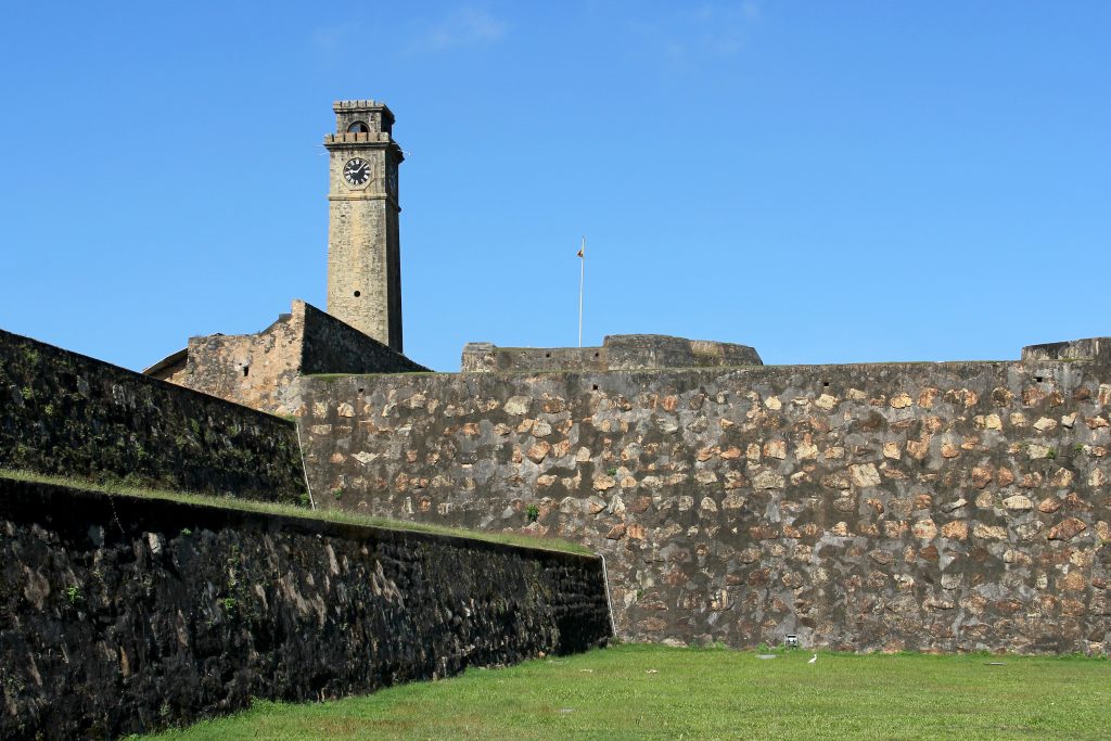 Stone clock tower rising above the massive land-side walls of Galle Fort, with a lawn in front