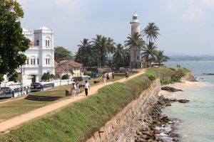 People walking the grassy rampart of Galle Fort above the sea wall, with the white Meeran Mosque and the lighthouse ahead