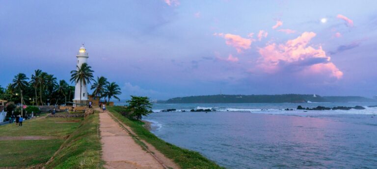 Galle Lighthouse and the grassy fort ramparts at dusk, with the sea and a wooded headland across the bay