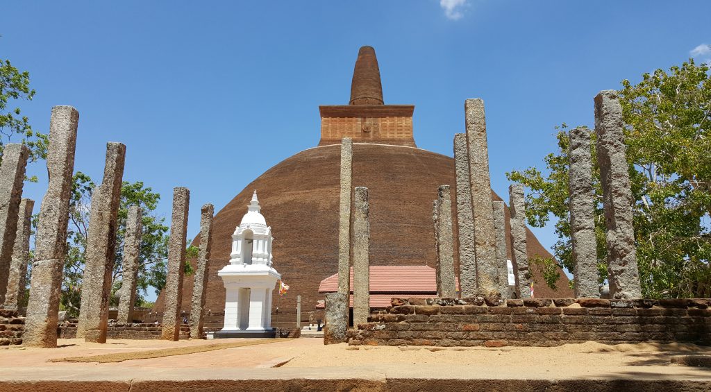 The brown brick Abhayagiri stupa rising behind rows of ancient stone pillars and a small white shrine, under a clear blue sky
