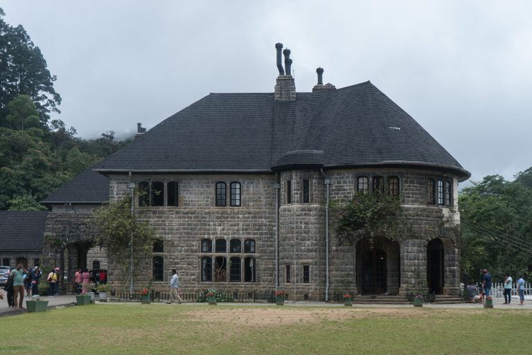 The grey stone front of Adisham Bungalow with its steep slate roof and round tower, behind a lawn