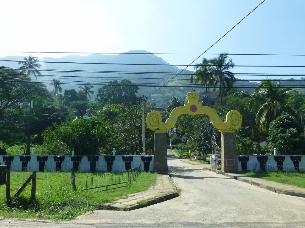 The arched entrance to Aluvihara beside the main road, with a lane leading in between white walls and forested hills behind