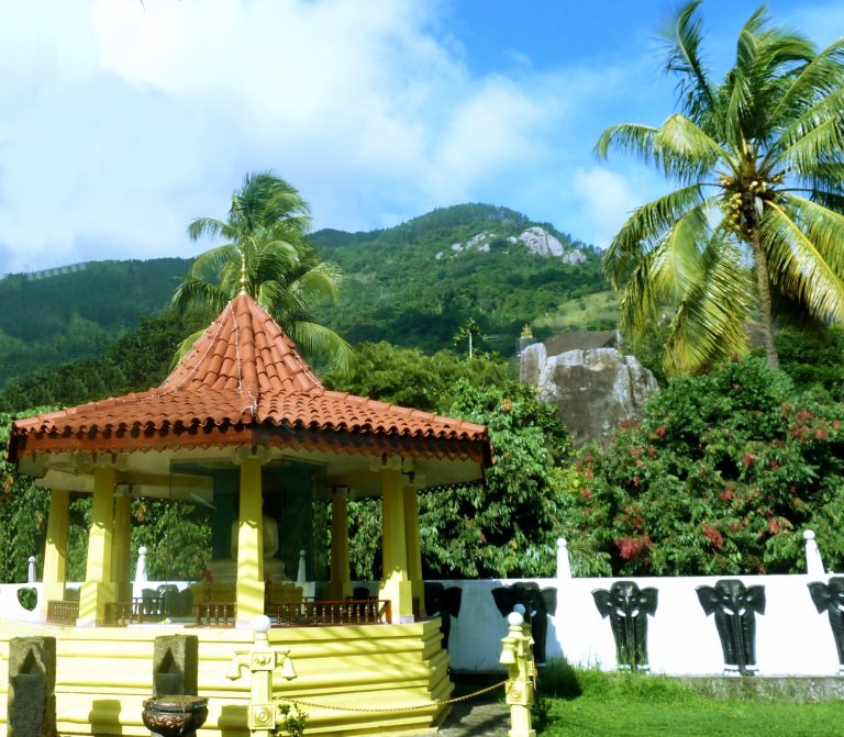 A tiled pavilion with a seated Buddha in front of the large boulder of Aluvihara, with a small shrine on top of the rock and forested hills behind