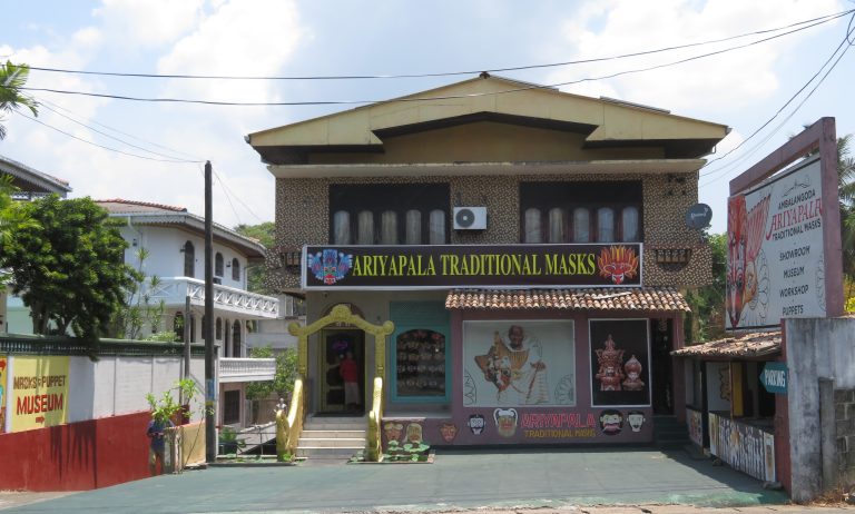 The front of the Ariyapala Traditional Masks building in Ambalangoda, with painted masks on the facade and a roadside sign for the showroom, museum, workshop and puppets