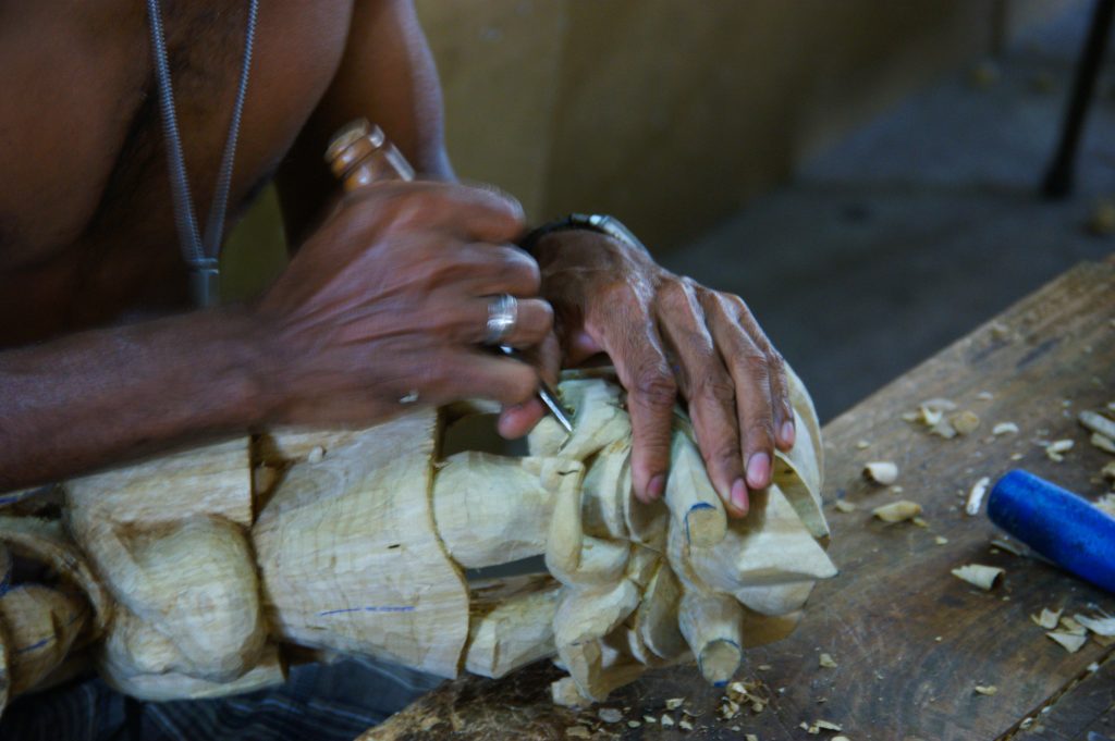 A carver's hands shaping an unpainted wooden mask with a small knife at a workbench scattered with wood shavings