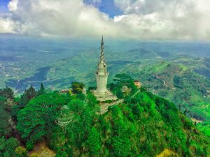 Aerial view of the white spiral Ambuluwawa Tower on a forested hilltop, with green hills and valleys stretching away under cloud