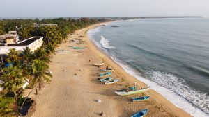 Aerial view of Arugam Bay beach in early light, with fishing boats on the sand, palms and guesthouses on the left and waves breaking along the shore