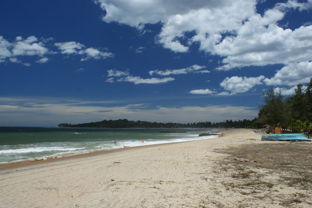A long, pale beach at Arugam Bay with waves rolling in on the left, an upturned boat at the back of the sand and a wooded point in the distance