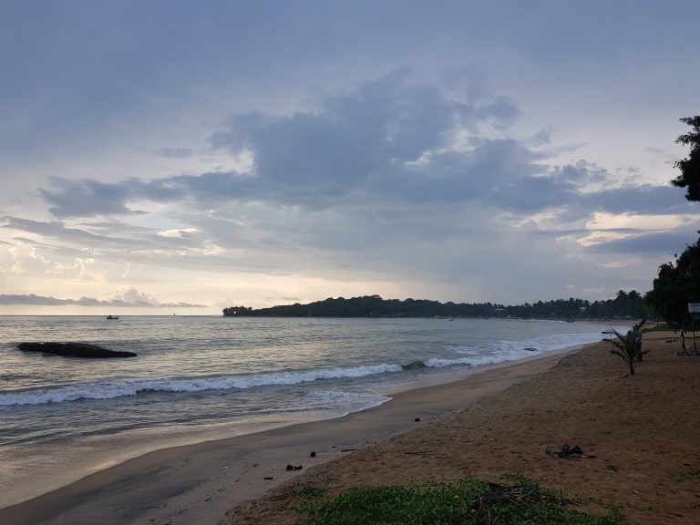 Waves breaking along the sand at Arugam Bay at dusk, with a dark rock in the surf and the wooded point in the distance