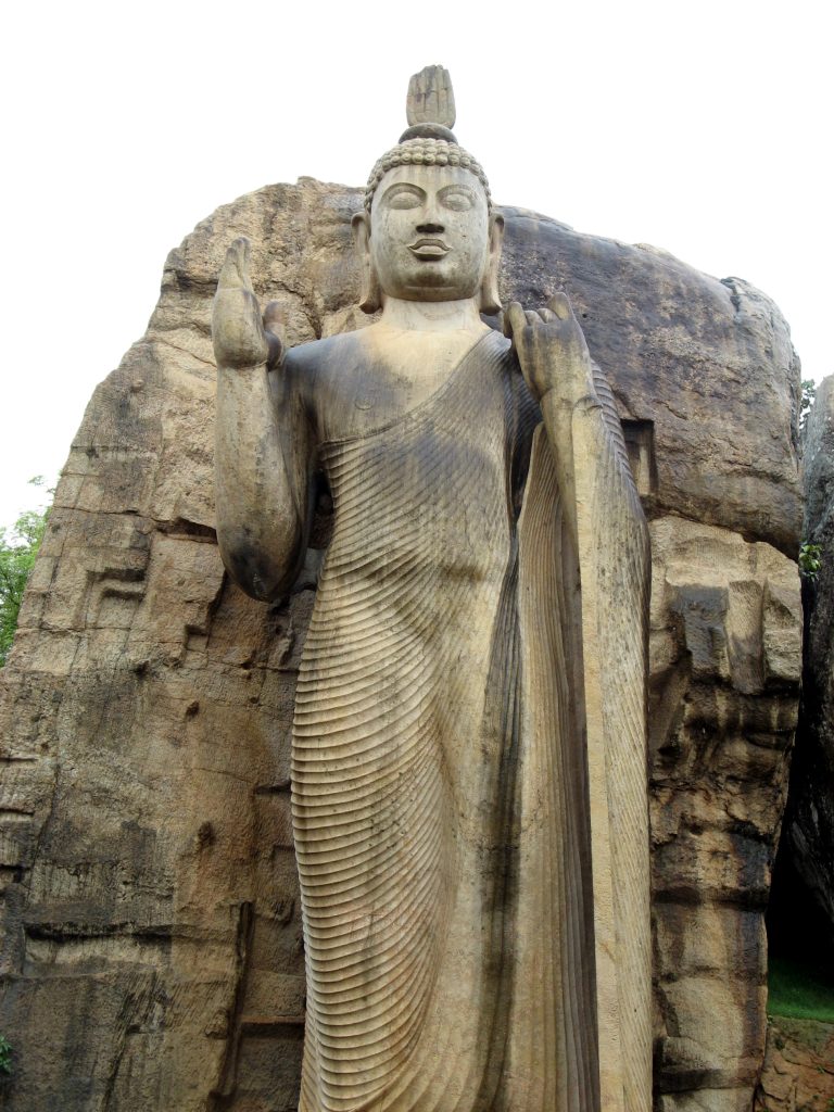 Close view of the Avukana Buddha from below, showing the raised right hand, the left hand holding the robe at the shoulder and the finely pleated robe