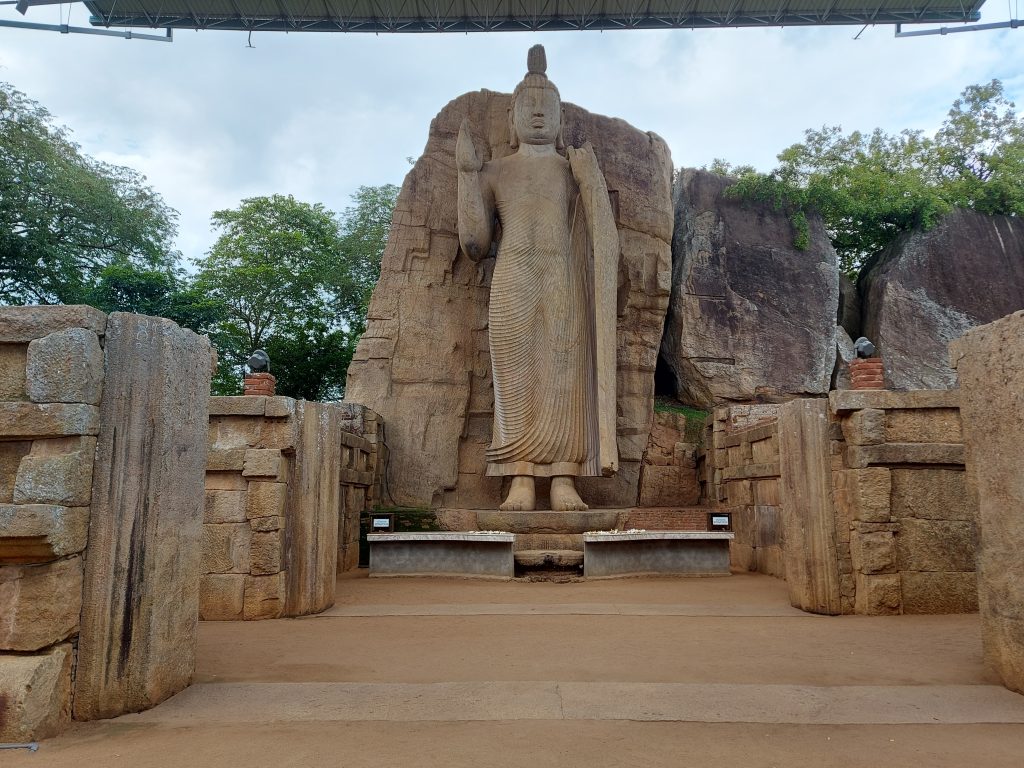The Avukana standing Buddha carved against a granite rock face, right hand raised, under a metal roof, framed by the low stone walls of the old image house