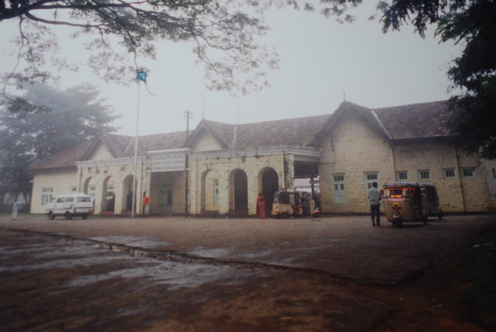 The long cream-coloured colonial building of Badulla railway station with arched entrances and a tiled roof, tuk-tuks parked in front on a misty day