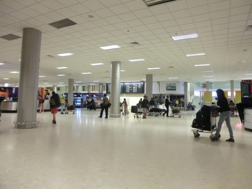 Travellers with luggage trolleys in the arrivals concourse at Bandaranaike International Airport, with a Mobitel counter and flight information screens behind