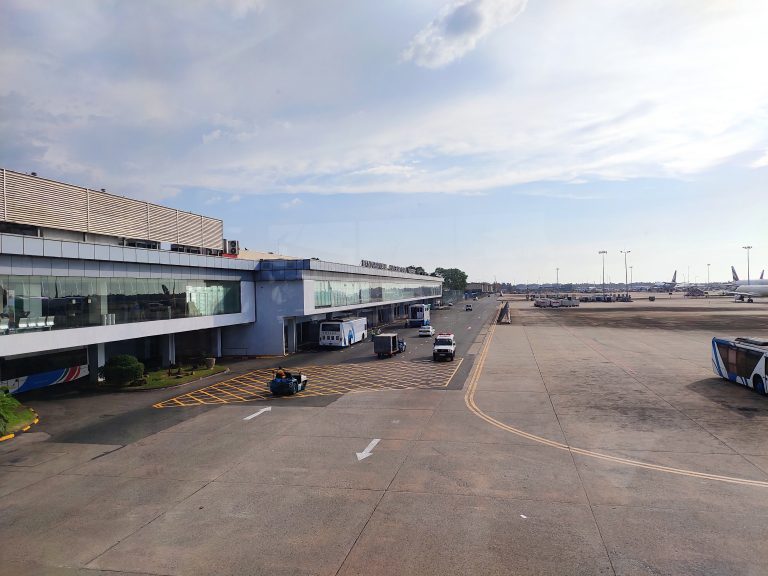 The terminal of Bandaranaike International Airport with its name on the roof, seen across the apron with airport buses and service vehicles