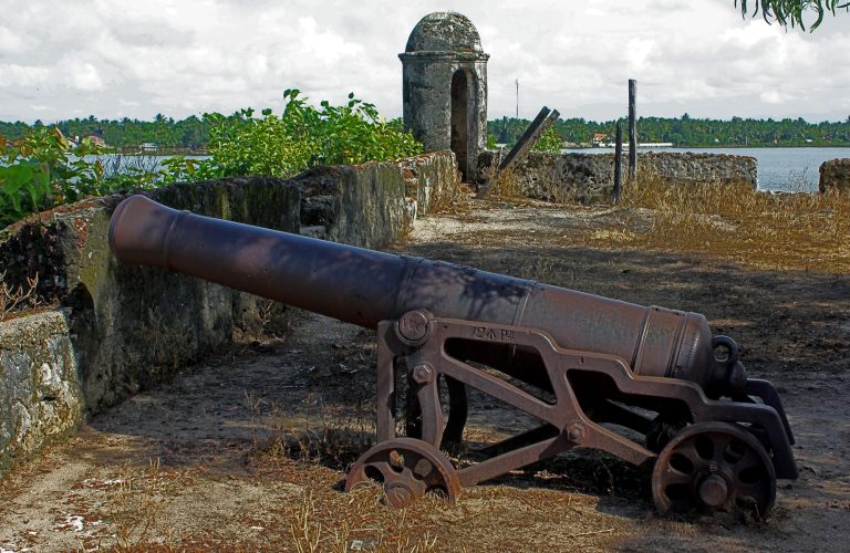An old iron cannon on a wheeled carriage on the rampart of Batticaloa Fort, with a domed stone sentry box behind and the lagoon and palm-lined shore beyond