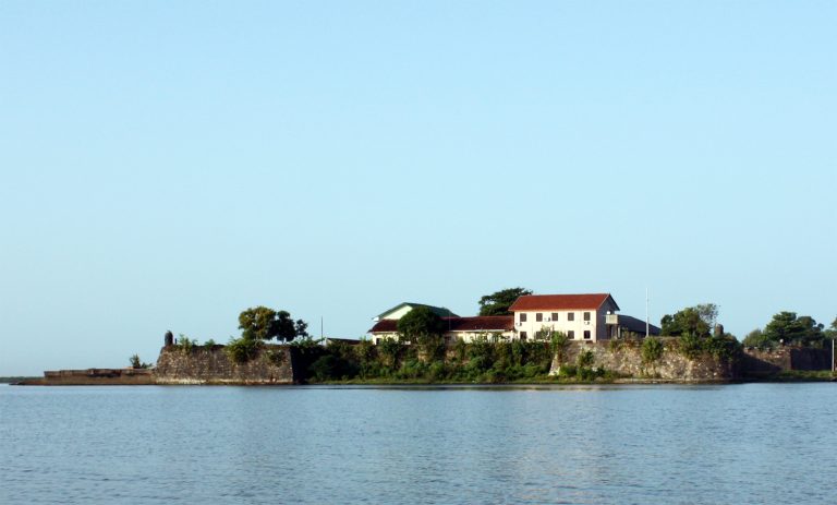 Batticaloa Fort seen across the calm lagoon, with low stone bastion walls and red-roofed buildings behind them