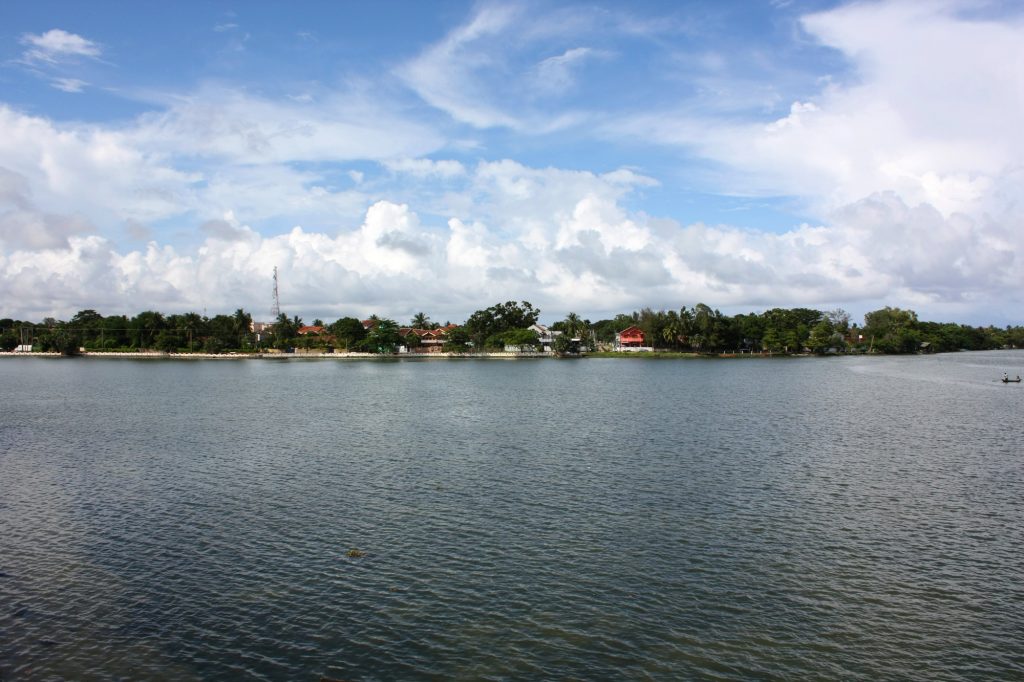 The wide Batticaloa Lagoon under a cloudy sky, with a line of houses and palm trees on the far shore and a small boat on the water