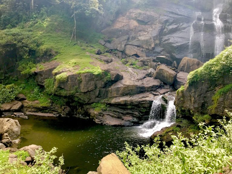 A green pool and small cascade over rocks below the main rock face of the falls, with mossy boulders around