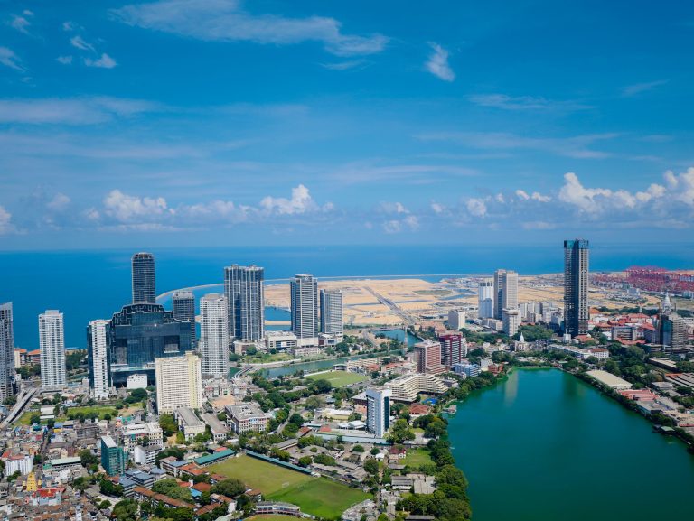 Aerial view of central Colombo: Beira Lake in the foreground, a cluster of high-rise towers beyond, and the sandy reclaimed land of Port City, the harbour cranes and the Indian Ocean behind