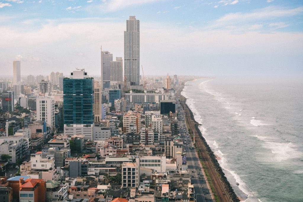Aerial view of Colombo's seafront south of the centre, with dense buildings and tall towers on the left and a railway line and road running beside the waves on the right