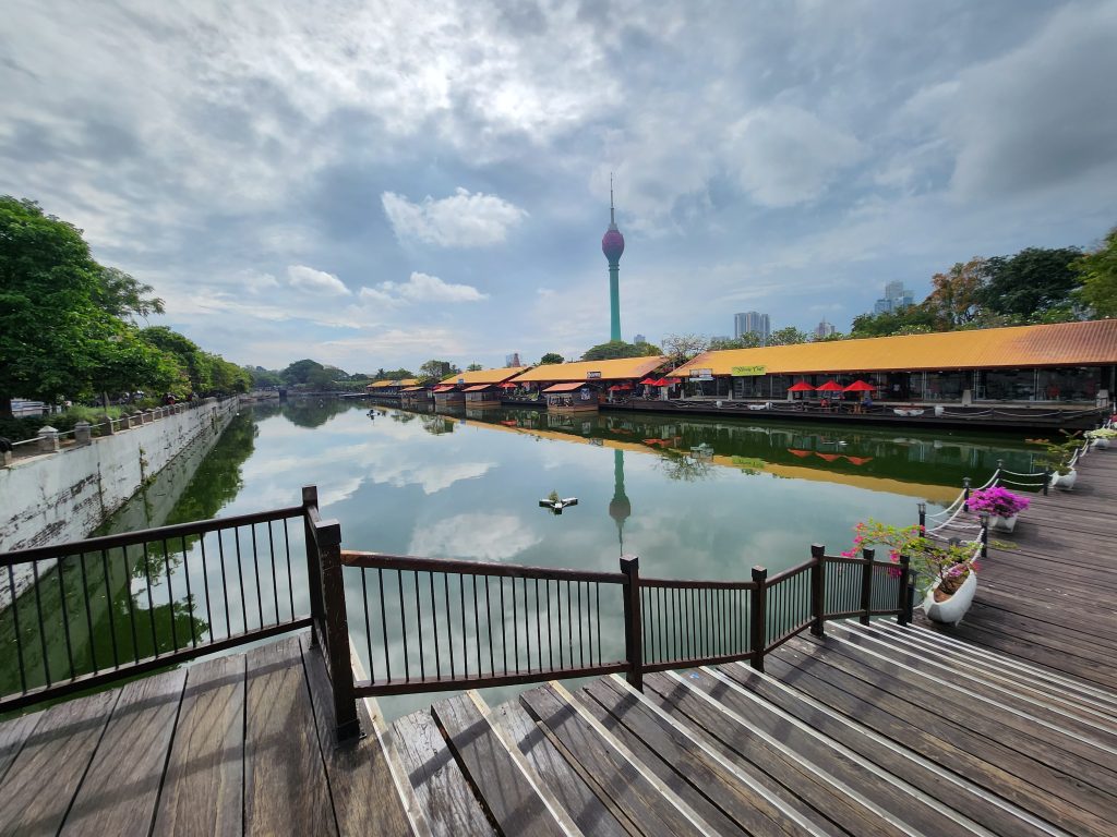 The Colombo Lotus Tower rising above Beira Lake, with the lakeside market buildings and a wooden walkway in the foreground
