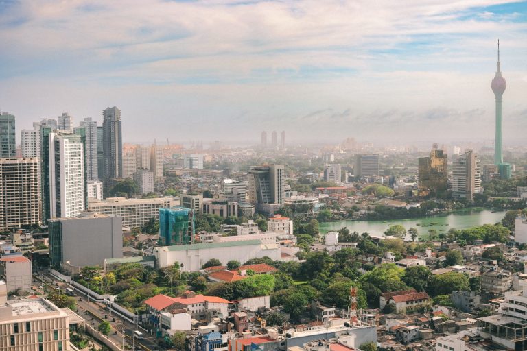 View over Colombo's rooftops, trees and apartment towers towards Beira Lake, with the green-and-pink Lotus Tower rising on the right