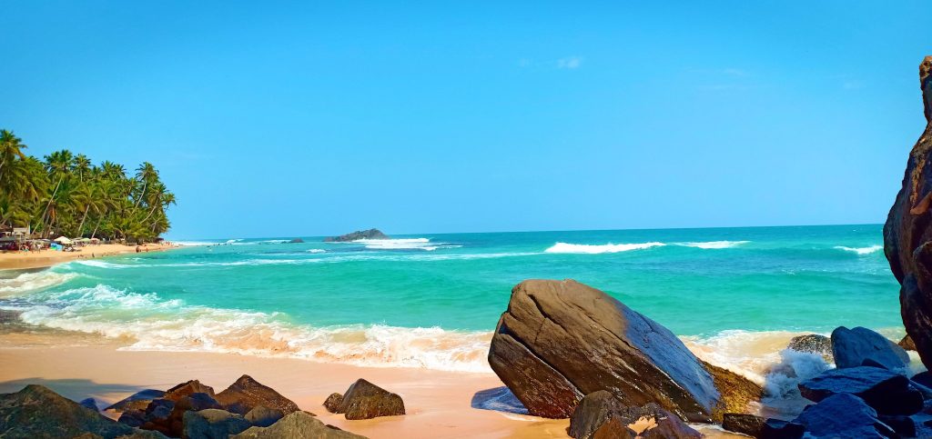 A pale sandy beach at Dalawella with waves rolling in over turquoise water, large boulders in the foreground and a palm-lined shore on the left