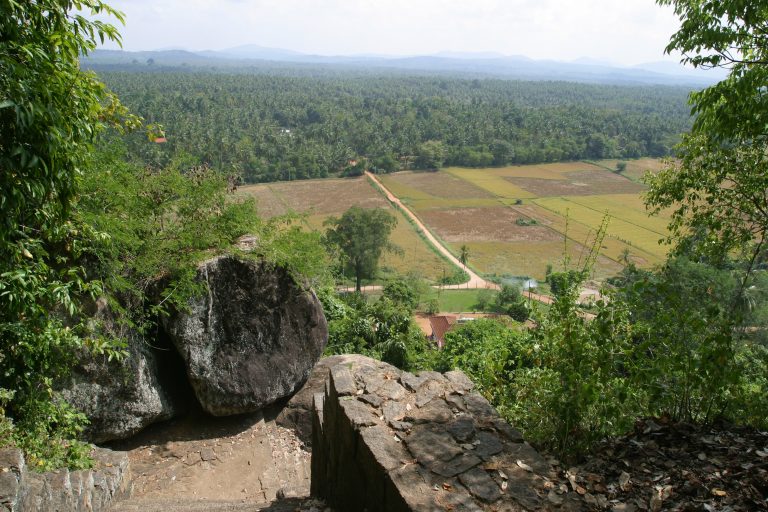 Old stone stairway and wall beside a large boulder near the top of the Dambadeniya rock, looking down over paddy fields and coconut groves