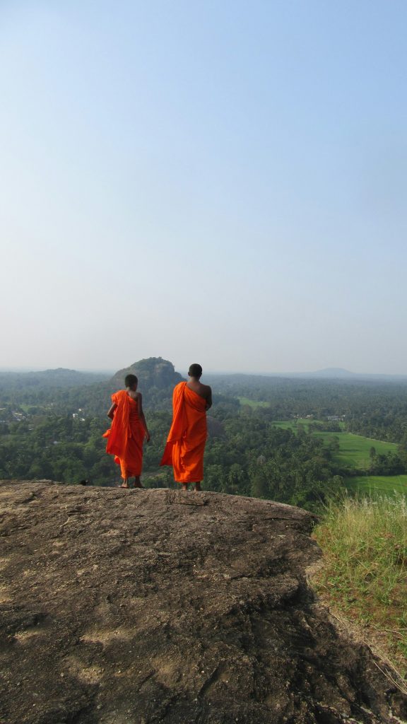 Two young Buddhist monks in orange robes on the bare rock summit of Dambadeniya, looking out over forest, paddy fields and a nearby rock hill