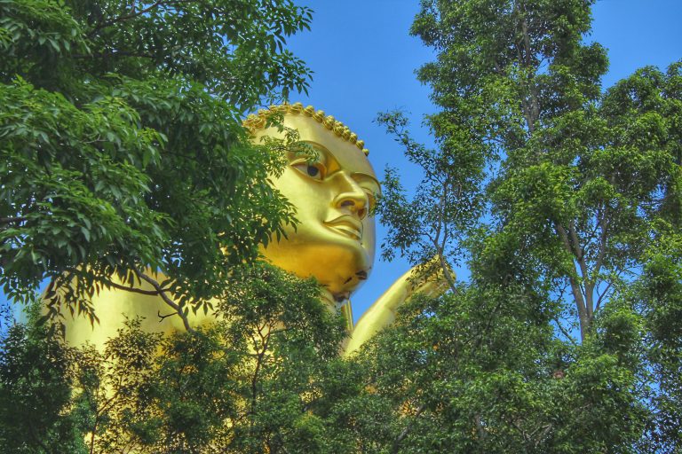 The head and raised hand of the giant golden Buddha statue at Dambulla, half framed by green trees against a blue sky