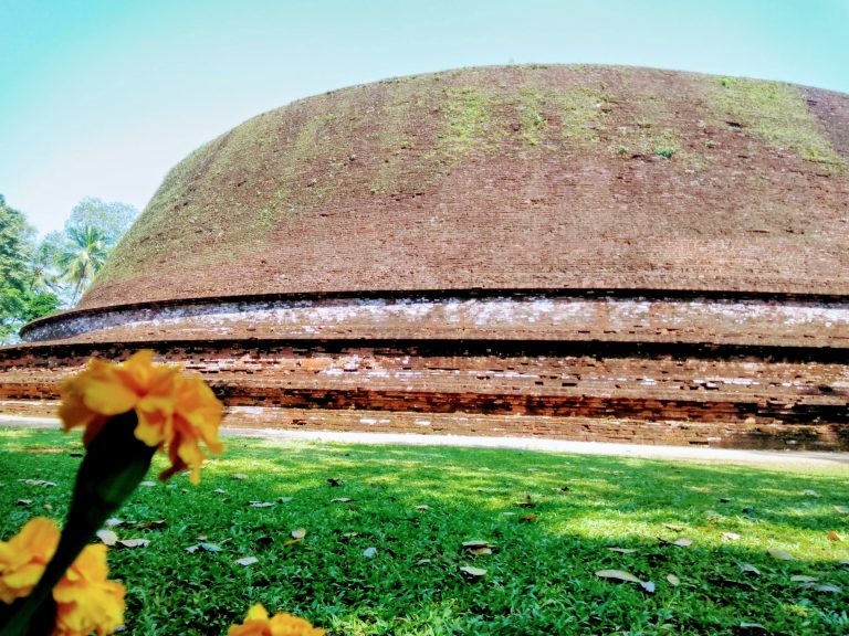 The low, flat-topped brick dome of Dedigama Kota Vehera on banded brick terraces, with moss on its slopes and a green lawn and marigolds in front