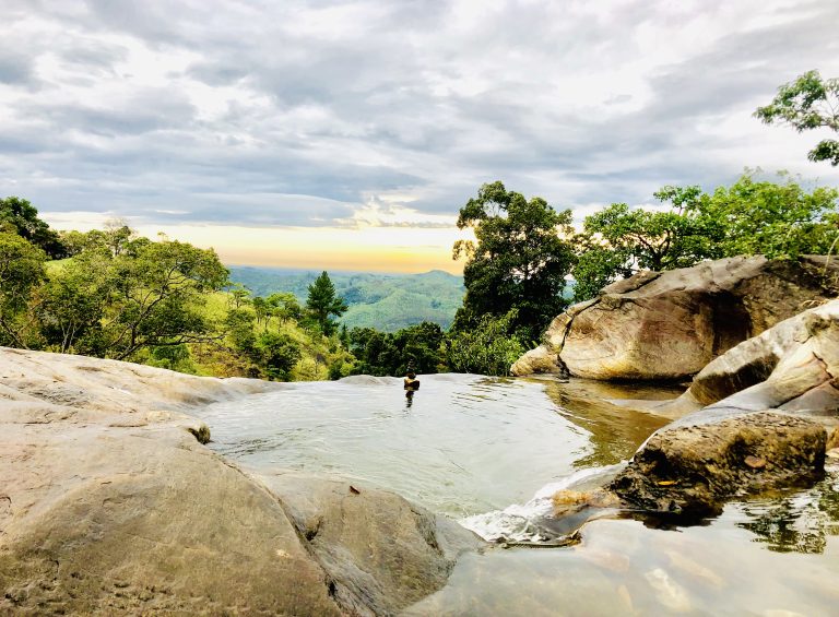A still rock pool on smooth boulders above Diyaluma Falls, with forested hills and a cloudy evening sky beyond
