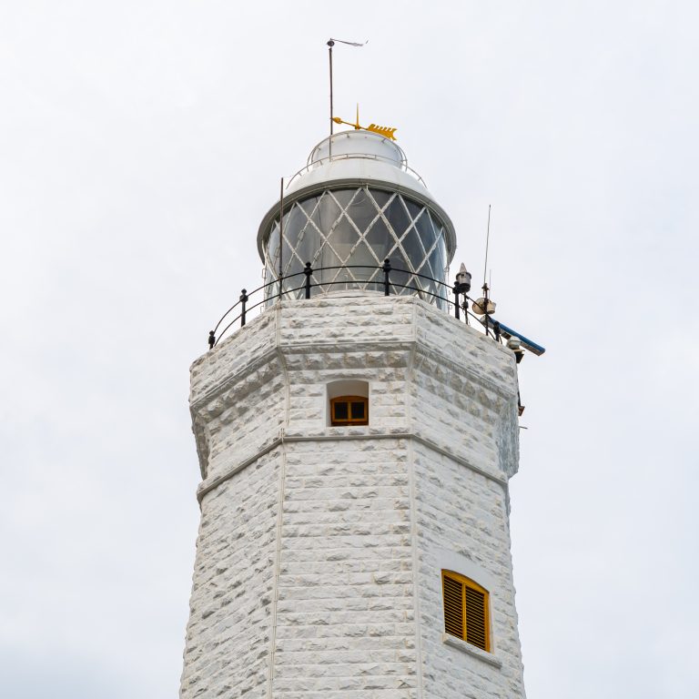 The top of Dondra Head Lighthouse: white octagonal stonework, yellow-shuttered windows, a railed gallery and the glazed lantern with a weather vane