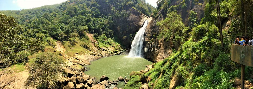Wide view of Dunhinda Falls dropping into a green pool ringed by boulders and forest, with visitors at a viewing platform on the right