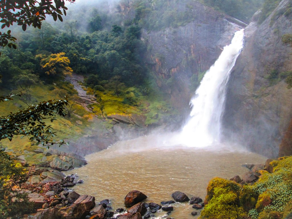 Dunhinda Falls pouring into a brown pool in a rocky gorge, with spray rising and mist over the forested slopes