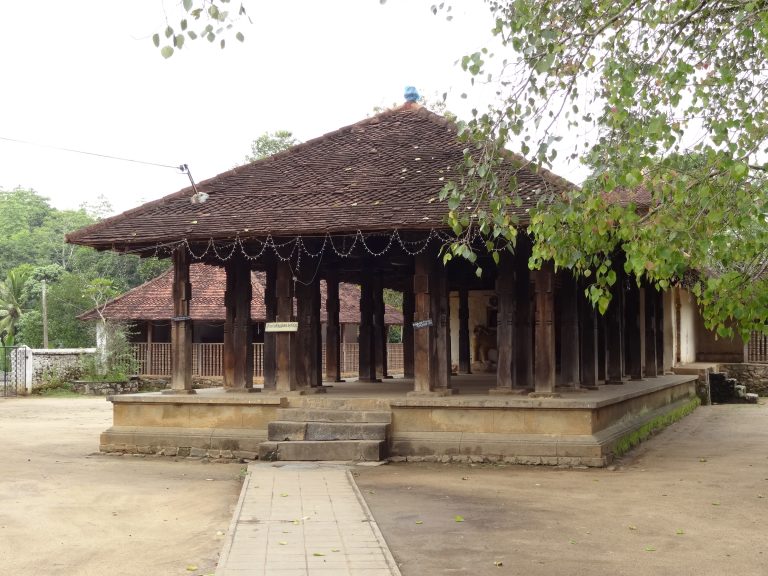 The open wooden hall of Embekke Devalaya, rows of dark carved pillars under a tiled hipped roof, on a raised stone base in a sandy courtyard