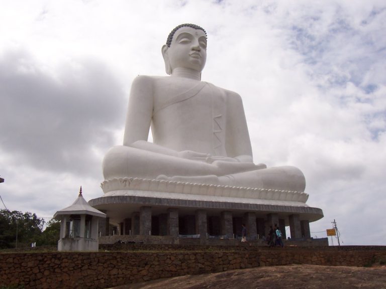 Giant white seated Buddha on a pillared base on the bare granite summit of Ethagala, with a small white shrine to the left and a cloudy sky
