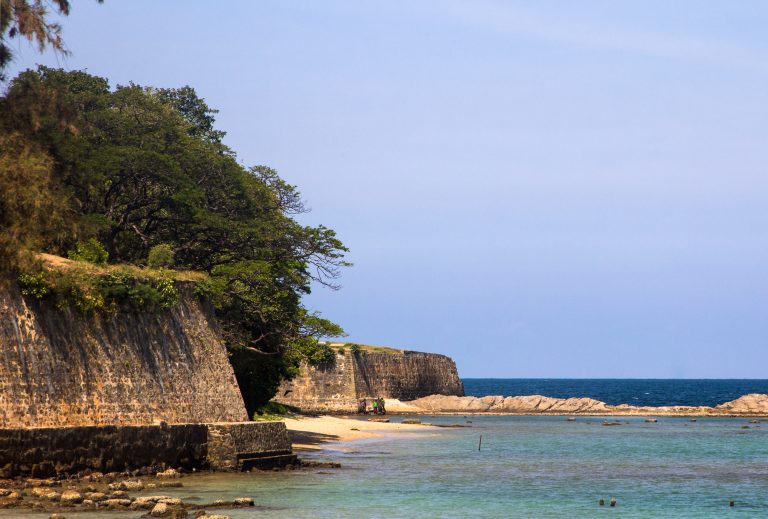 Old stone ramparts of Fort Frederick rising above a small beach, with clear shallow water and the open sea beyond