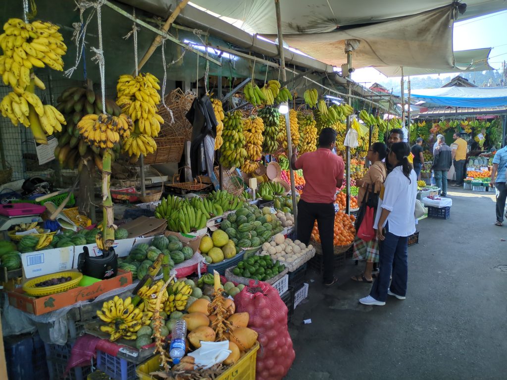Bunches of bananas, melons, mangoes and oranges piled on stalls under awnings at a fruit market, with shoppers in the aisle