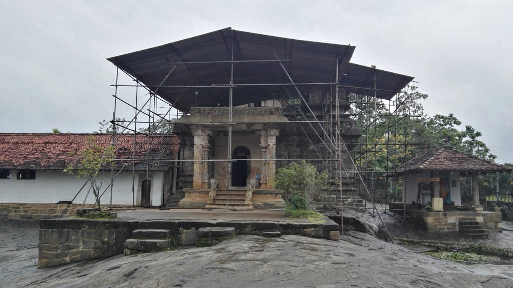 The grey stone image house of Gadaladeniya with a carved pillared porch, under a metal protective roof held up by scaffolding, on a sheet of bare rock