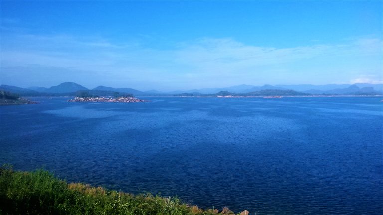The wide blue water of Senanayake Samudraya with small rocky islands and a line of hazy hills along the far shore