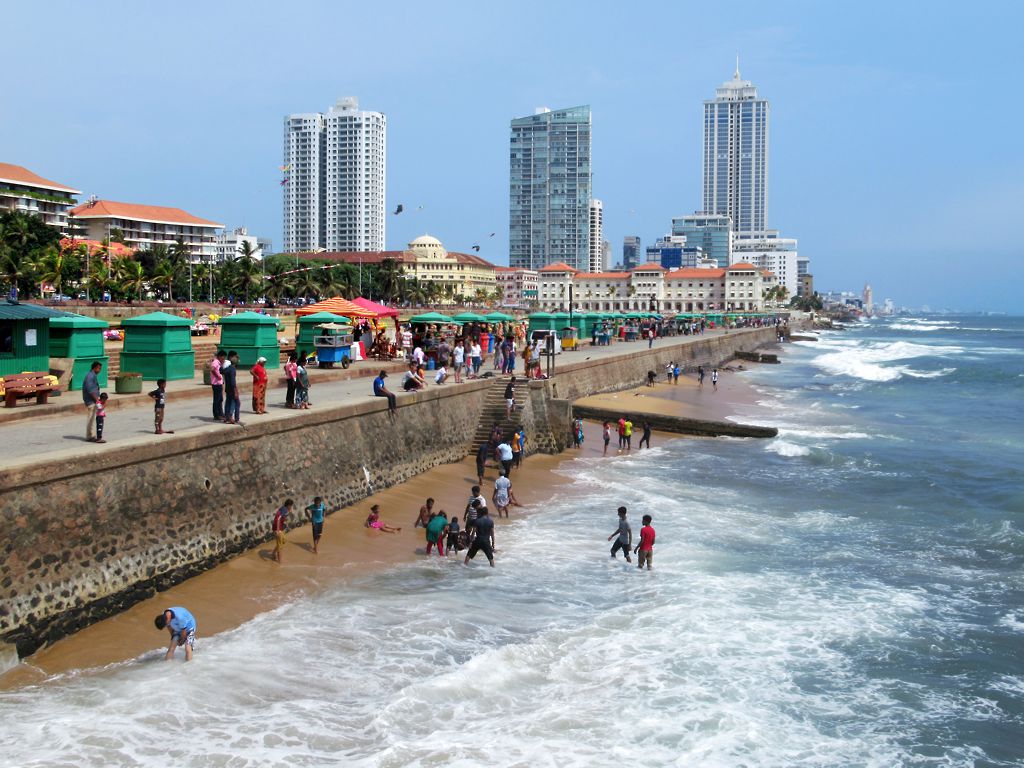 People paddling in the waves below the Galle Face sea wall, with kiosks along the promenade and the Galle Face Hotel and high-rise towers beyond