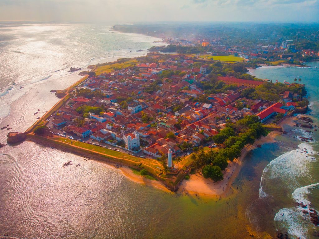 Aerial view of Galle Fort on its headland: red-roofed old town inside the ramparts, the white lighthouse at the tip, a small beach and the sea on three sides