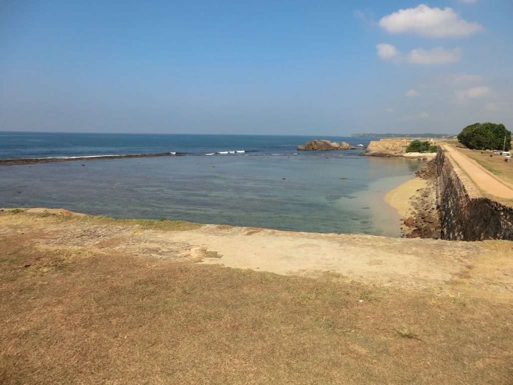 The wide, sun-baked grass top of Galle Fort's western rampart, with no railing along the edge and the sea and a rocky reef beyond