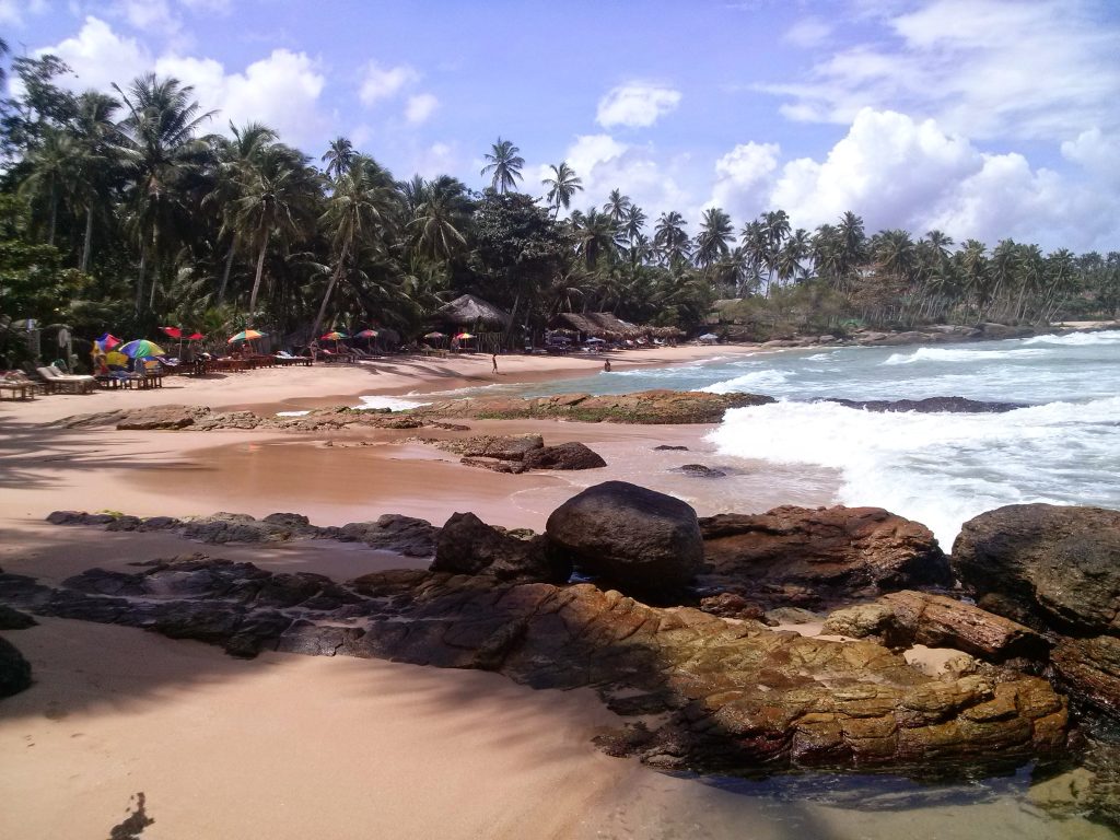 Goyambokka beach, a small sandy cove with dark rocks in the foreground, waves and palm-shaded beach huts
