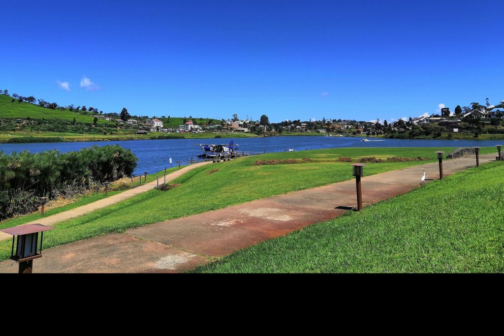Gregory Lake in Nuwara Eliya seen across the water from a lakeside path, with town buildings and tea-covered hills on the far shore