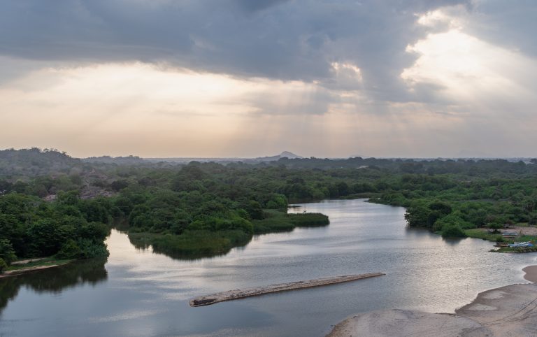 The Heda Oya river winding through green scrub and forest towards the coast under evening cloud, with a sandbank in the foreground
