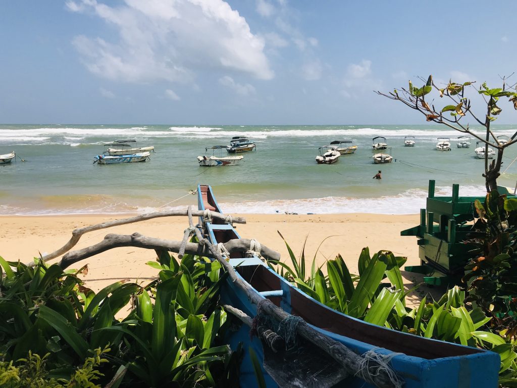 Small canopied tour boats moored in the calm lagoon inside Hikkaduwa's reef, with waves breaking on the reef edge beyond and a wooden outrigger canoe on the sand