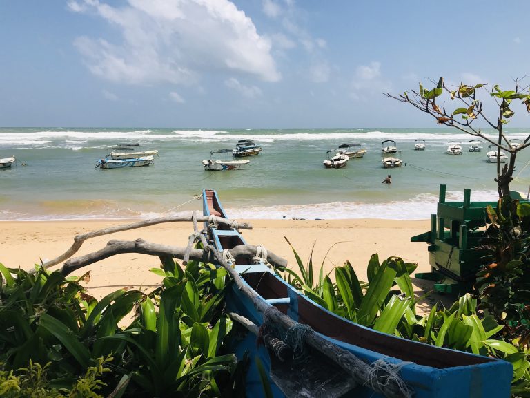 Small canopied tour boats moored in the calm lagoon inside Hikkaduwa's reef, with waves breaking on the reef edge beyond and a wooden outrigger canoe on the sand